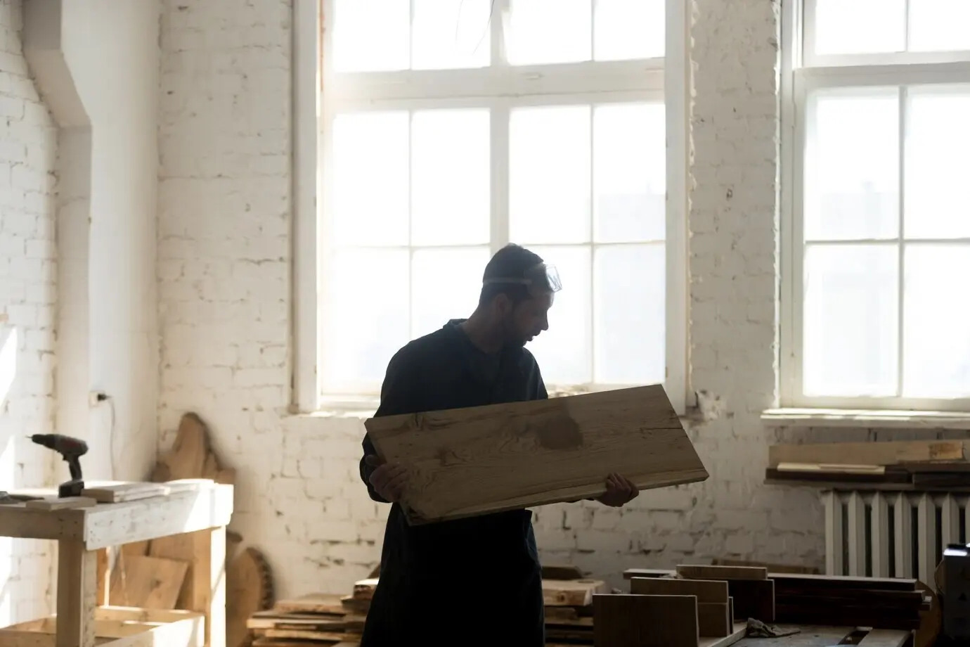 Joven carpintero sosteniendo una tabla de madera, trabajando la madera en el interior de un taller de carpintería.